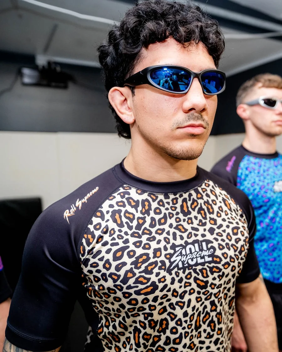 Someone in wraparound sunglasses wearing the Classic Pat leopard rash guard, with a teammate in Psychedelic Pat behind.